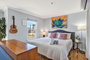 A bedroom with a bed, a guitar, a window, a painting, and a bedside table.