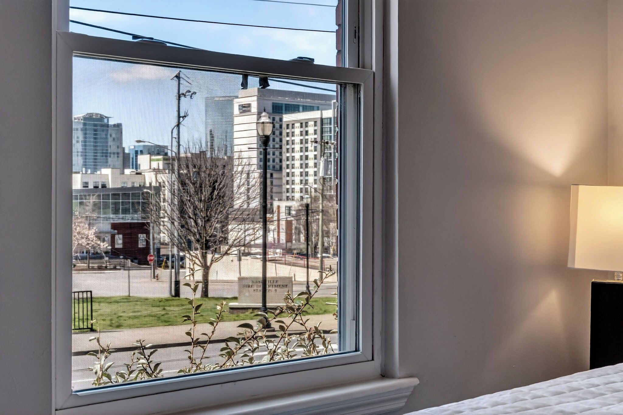 A hotel room with a large window, a bed, a bedside table, and a lamp.