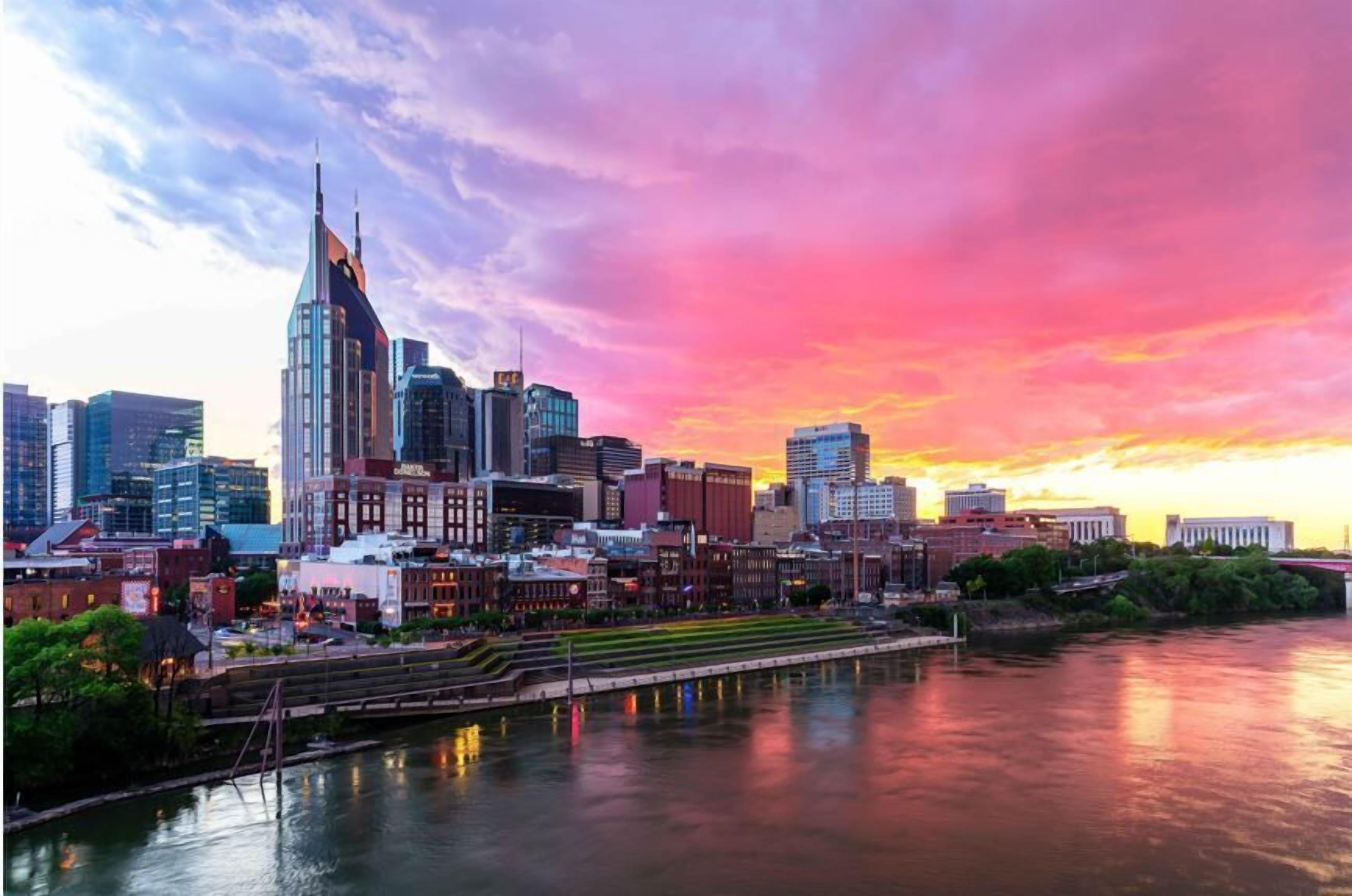 A city skyline at sunset with modern buildings and a river in the foreground.