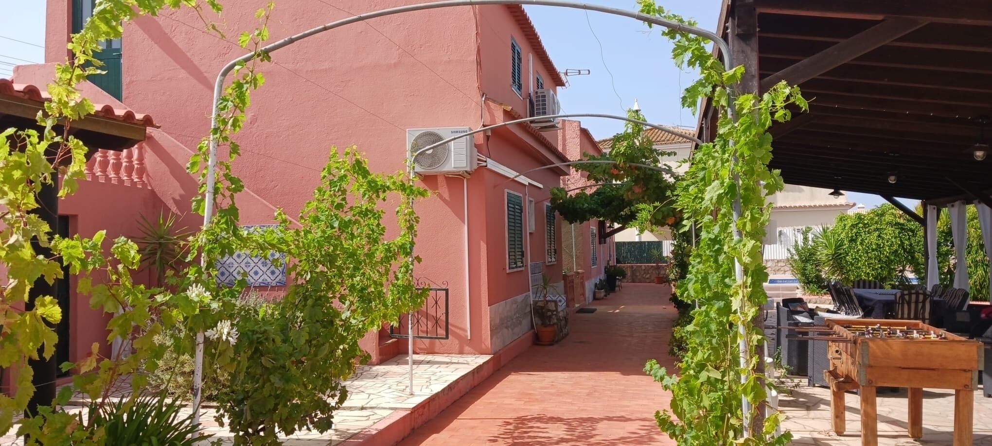 Un vicolo stretto con un edificio rosso, una zona salotto esterna coperta e una pergola ricoperta di edera.