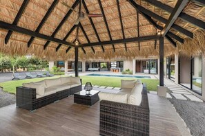 An outdoor lounge area with a thatched roof, wicker furniture, and a pool in the background.