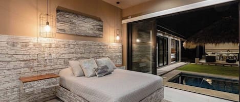 A bedroom with a large bed, a ceiling fan, a stone wall, and a view of a pool.