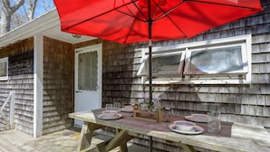 Una terraza de madera con una mesa de picnic dispuesta para cuatro personas, un paraguas rojo y una ventana con contraventanas.