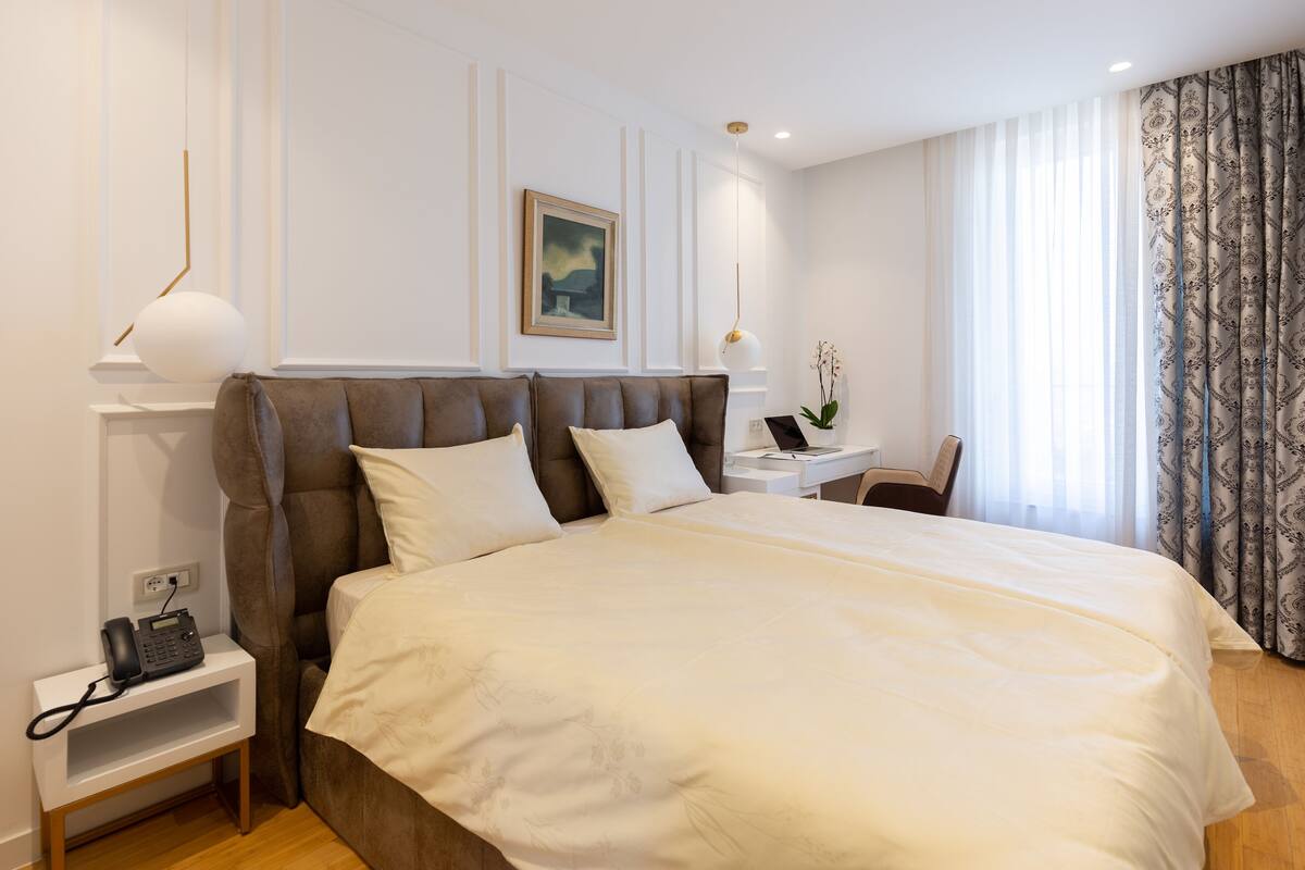 Een moderne hotelkamer met een groot bed, nachtkastje, lamp en uitzicht op het buitenland door dunne gordijnen.