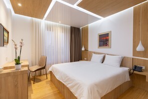 A modern hotel room with a large bed, a desk with a plant, a chair, and a painting on the wall.