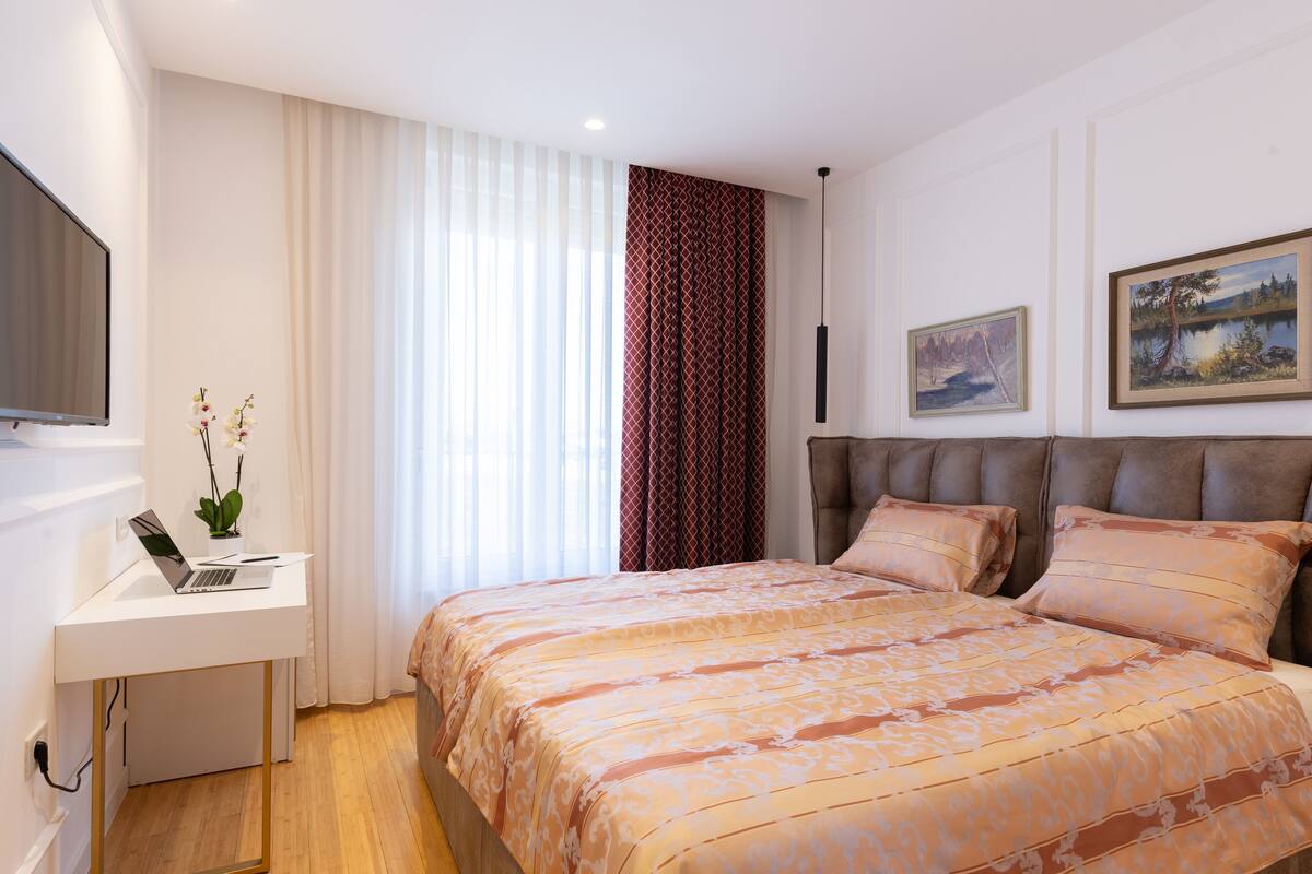 Een slaapkamer met een bed, een bureau met een laptop, een televisie en twee schilderijen aan de muur.