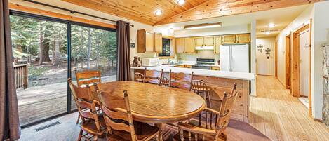 A wooden interior with a dining area, kitchen, and large windows overlooking a forest.