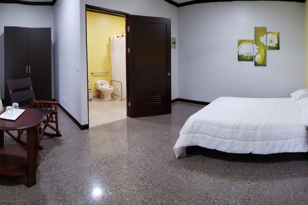 A hotel room with a bed, bedside table, chair, and a small table with a chair. There is a bathroom visible through an open door.