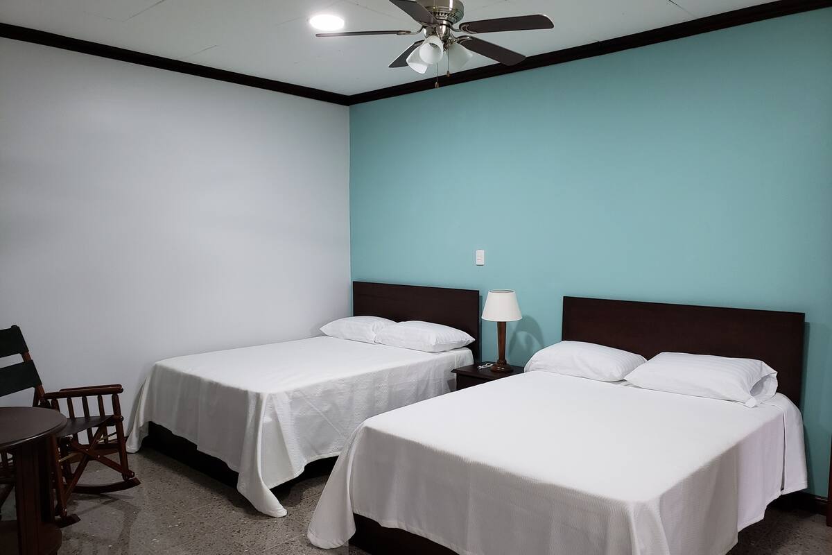 A hotel room with two single beds, a desk, a chair, a nightstand with a lamp, and a ceiling fan.