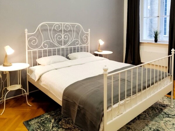 Un lit bien fait, avec une structure en métal blanc, une couverture grise et deux oreillers blancs. On y trouve deux tables de chevet avec des lampes, une petite plante en pot sur le rebord de la fenêtre et un tapis à motifs sur le sol.