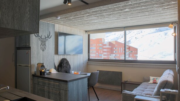 Una cocina moderna con refrigerador integrado, una zona de comedor con sofá y una mesa pequeña, y un ventanal con vistas a una montaña nevada y edificios.