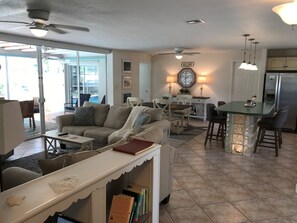 Un vaste espace de vie comprenant un canapé, une table basse et un petit coin bar avec des tabourets. On y trouve également une table à manger avec des chaises, une cuisine équipée d’un réfrigérateur et un ventilateur de plafond.