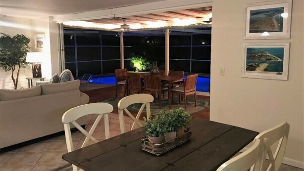 An indoor dining area with a dark wooden table, white chairs, and potted plants. There is a large window with a view of the night sky and a ceiling fan above. - Single open-concept home one block from the ocean with heated pool and lanai  (West Palm Beach)