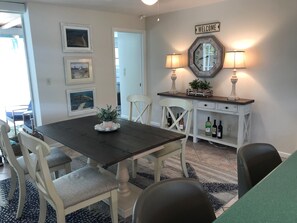 Un coin repas avec une table en bois foncé, des chaises blanches, une console blanche ornée d’une horloge et deux lampes fixées au mur.