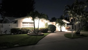 A residential house at night with a well-lit garage, a white picket fence, and a driveway leading to the front door. - Single open-concept home one block from the ocean with heated pool and lanai  (West Palm Beach)