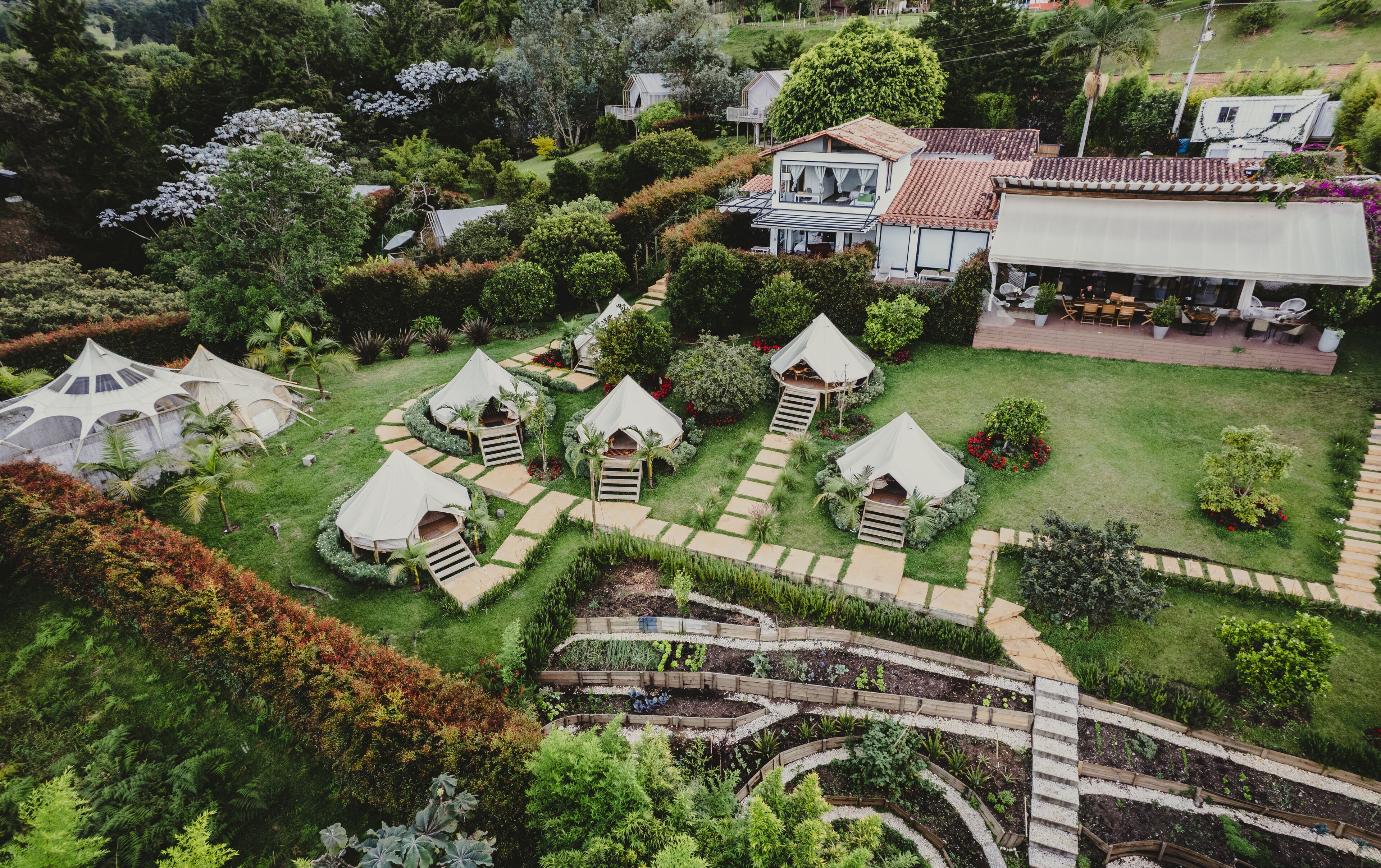 An aerial view of a resort with multiple bungalow-style tents arranged in a circular pattern around a central garden area, surrounded by lush greenery and a few residential buildings.