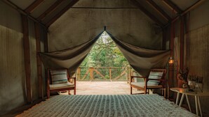 Een slaapkamer met een groot bed, twee stoelen, een klein tafeltje en uitzicht op een houten terras en bomen door gordijnen.