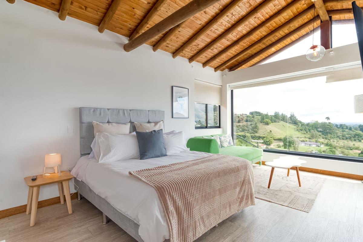 A bedroom with a bed, bedside table, lamp, and a view of a green landscape through large windows.