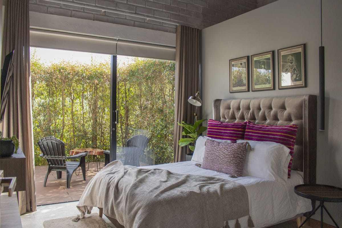 A bedroom with a large bed, a view of trees through the window, a small table, and a chair.