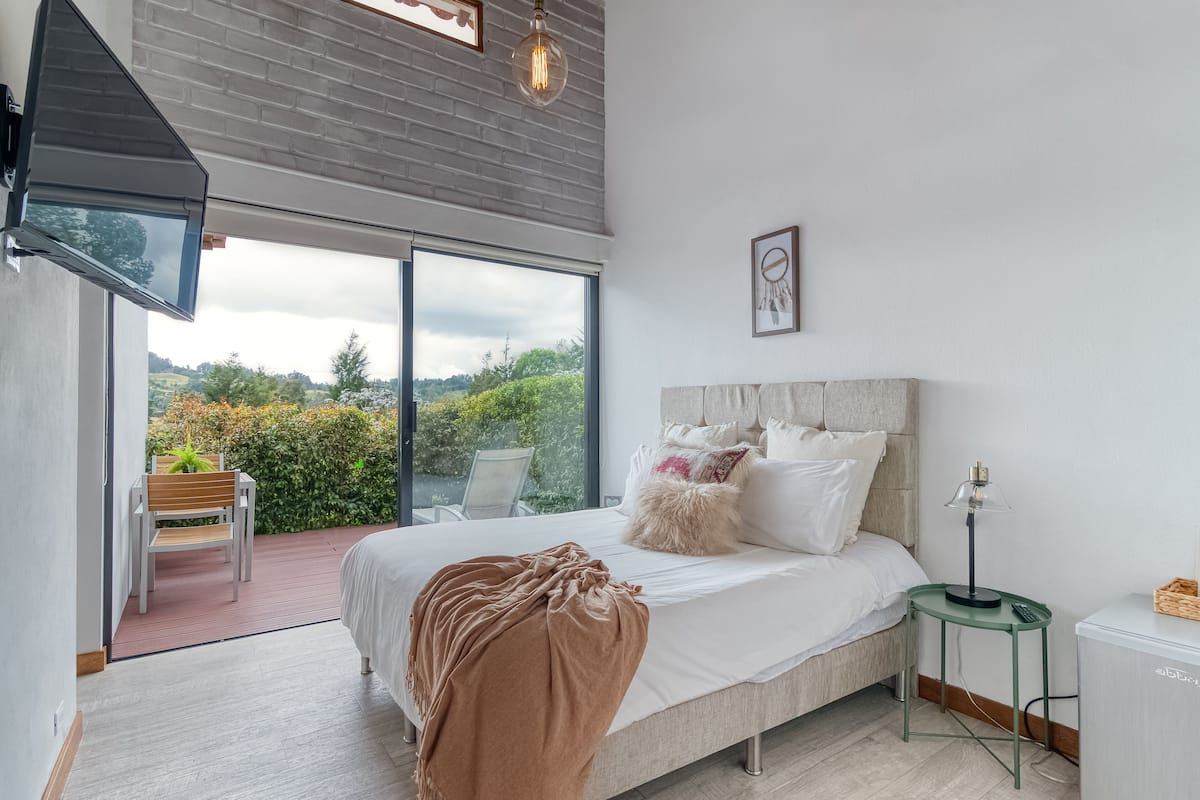 A bedroom with a large bed, a side table, a chair, and a view of greenery through a sliding glass door.