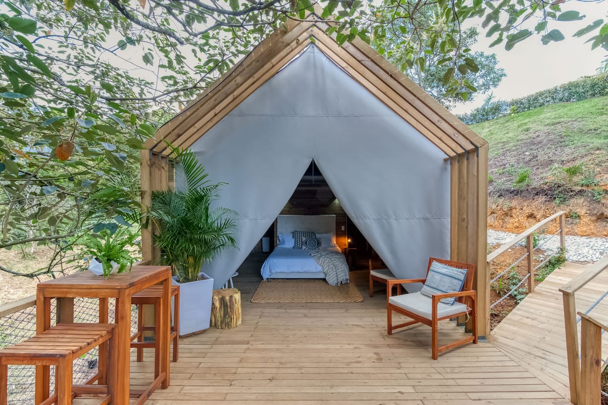 A cozy, tent-like structure with a bed, a chair, and a table on a wooden deck, surrounded by greenery.