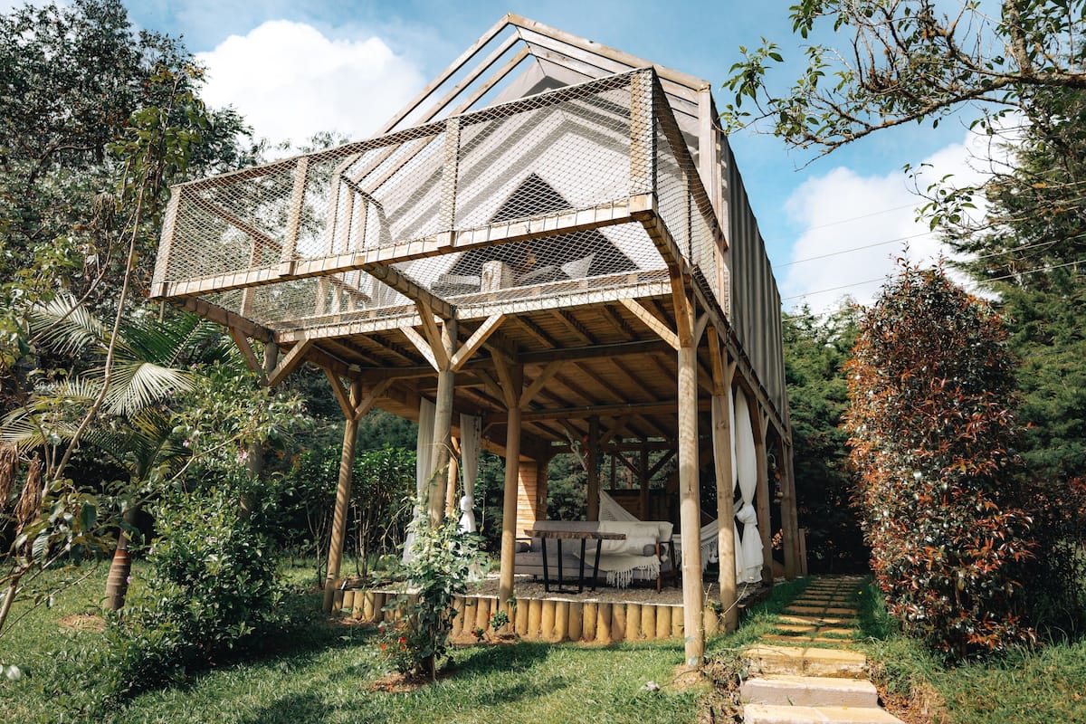 A wooden gazebo with a mesh roof, surrounded by lush greenery and a stone pathway.