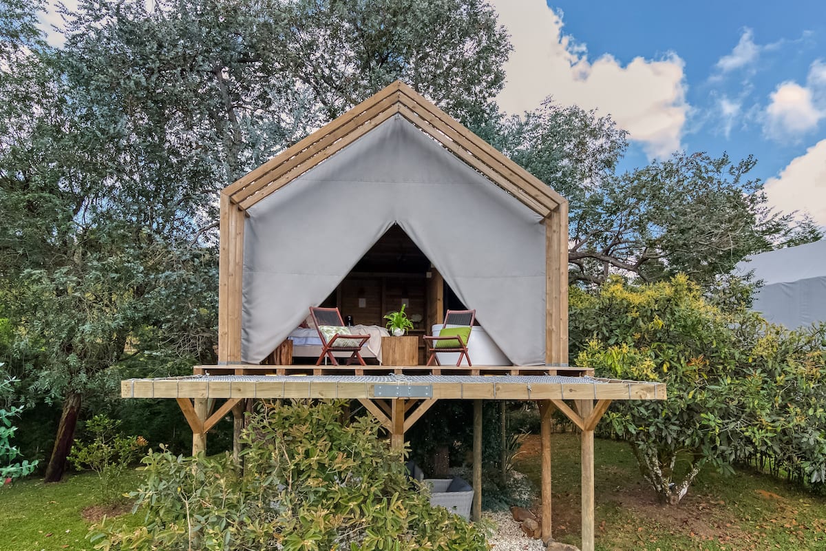 A wooden cabin with a triangular roof, a small balcony with two chairs and a table, surrounded by lush greenery.