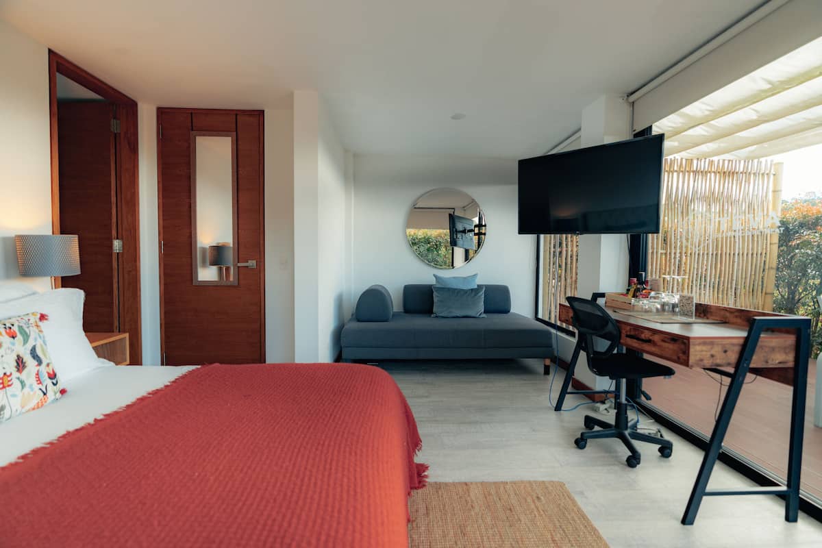 A modern hotel room with a bed, a desk, a sofa, a TV, and a large window.