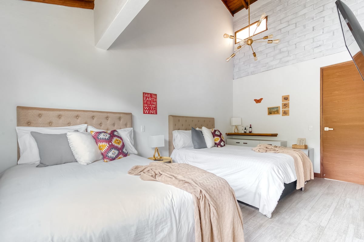 A bedroom with two beds, a wooden ceiling, a brick wall, a wooden door, and a small bedside table with a lamp.