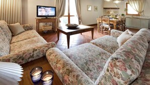 A living room with floral-patterned sofas, a television on a wooden stand, a dining table with chairs, and a kitchen area in the background. - Four leaf clover - Salvia (4 + 2) (Gambassi Terme)