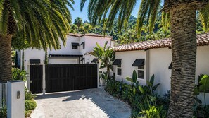 A two-story house with a tiled roof, a gated driveway, and lush tropical vegetation.