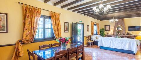 Un comedor con una mesa y sillas de madera, un jarrón de flores y una cama con cubrecama blanco al fondo.