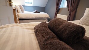 A bedroom with two beds, one with a white duvet and the other with a brown duvet. There is a window with curtains, a radiator, and a door with a brass handle. - Hideaway Cottage (Halesworth)