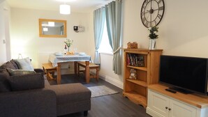 A living room with a sofa, a dining table with chairs, a TV on a wooden stand, a bookshelf, and a large wall clock. - Hideaway Cottage (Halesworth)