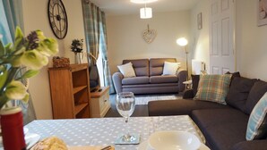 A cozy living room with a sofa, a coffee table, and a TV. There is a clock on the wall and a plant on the side. - Hideaway Cottage (Halesworth)