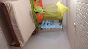 Une petite pièce étroite, carrelée, avec un lit recouvert d’un drap beige, un oreiller vert et orange, et un baldaquin jaune.