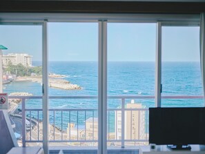Une vue à travers une fenêtre sur un paysage urbain côtier, avec des bâtiments, une route et la mer.