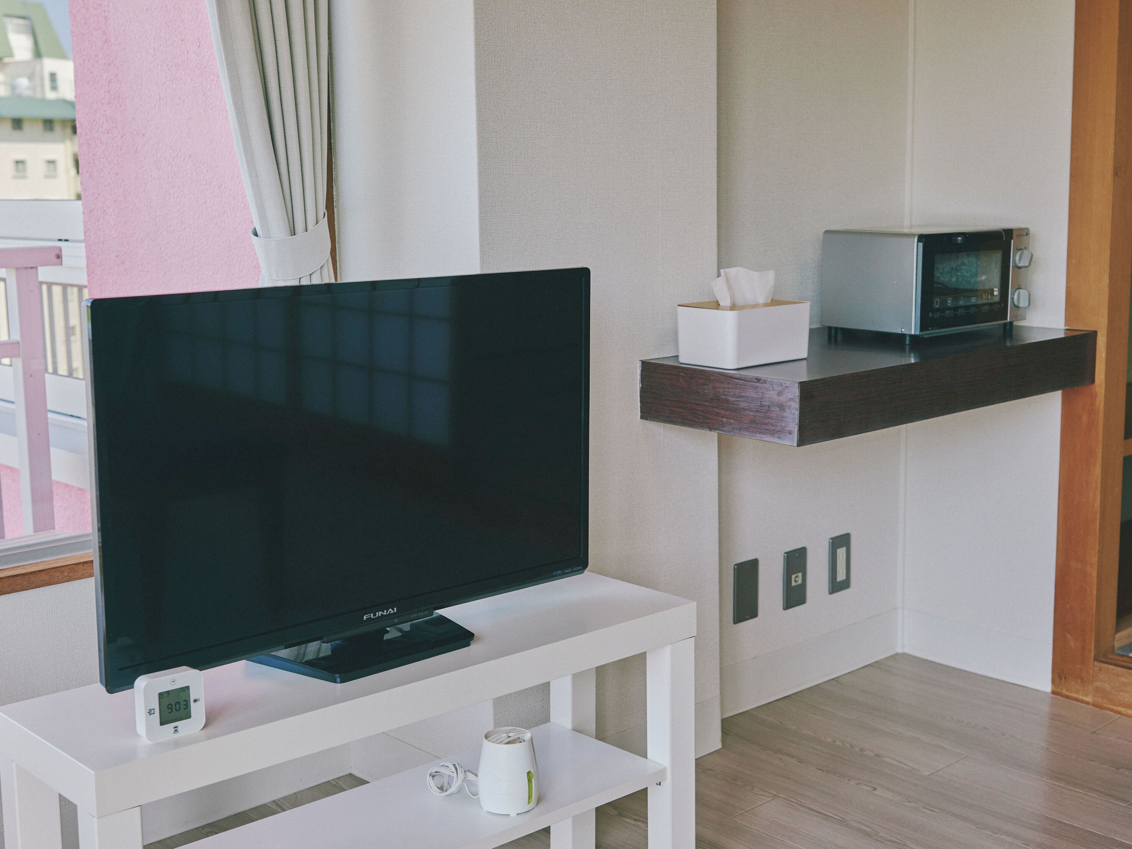 A modern living room with a flat-screen TV on a white stand, a microwave on a dark wooden shelf, and a small white table with a tissue box and a remote control.