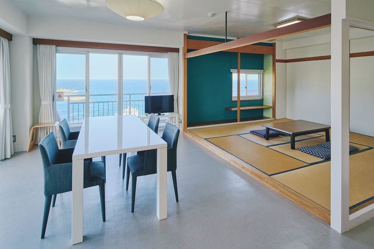 Ein modernes Hotelzimmer mit Esstisch, Stühlen, Fernseher, einer Couch, Tatami-Matten und großen Fenstern mit Meerblick.