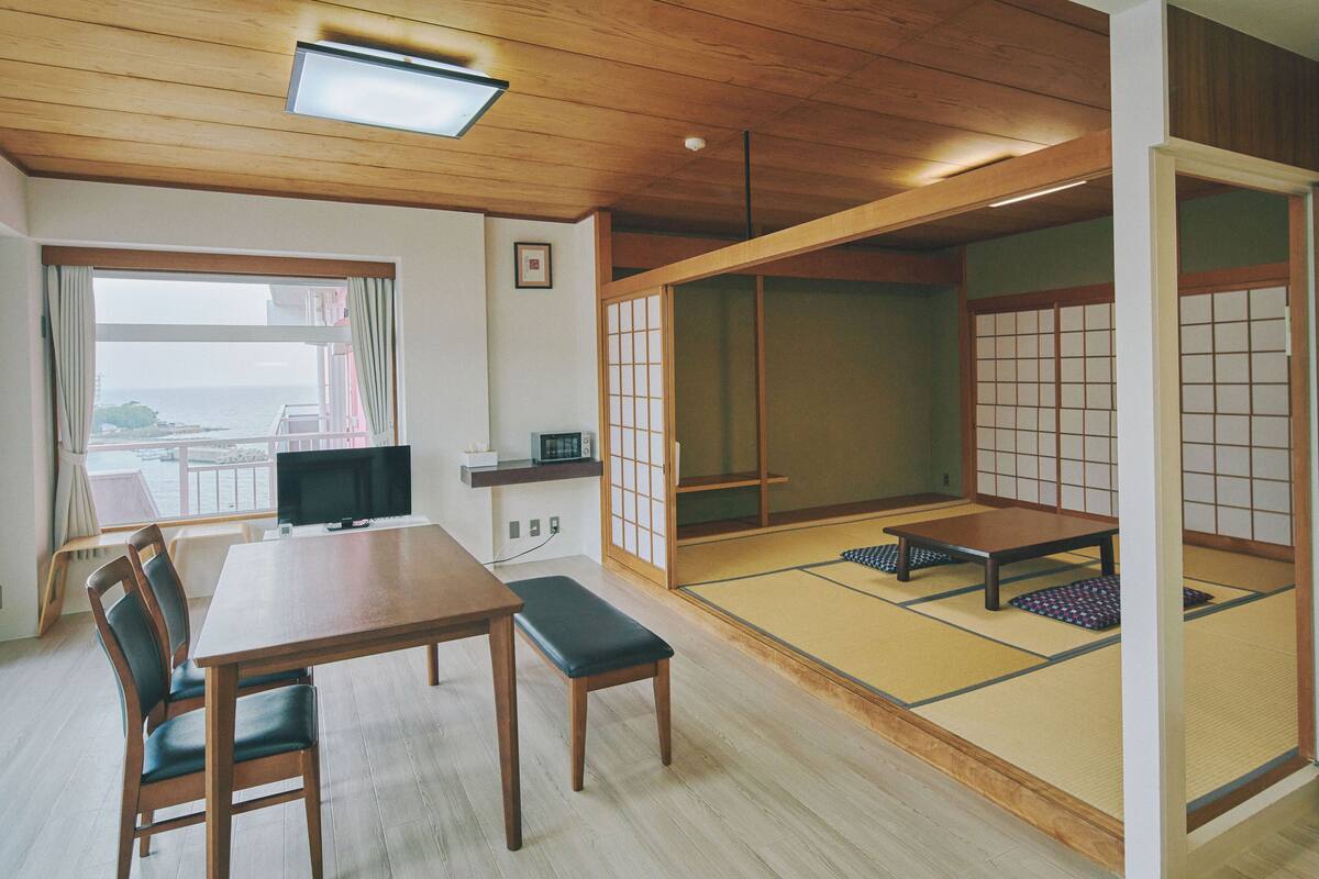 Ein traditioneller japanischer Raum mit Tatami-Boden, einem niedrigen Tisch, Stühlen, einem Fernseher und Meerblick.