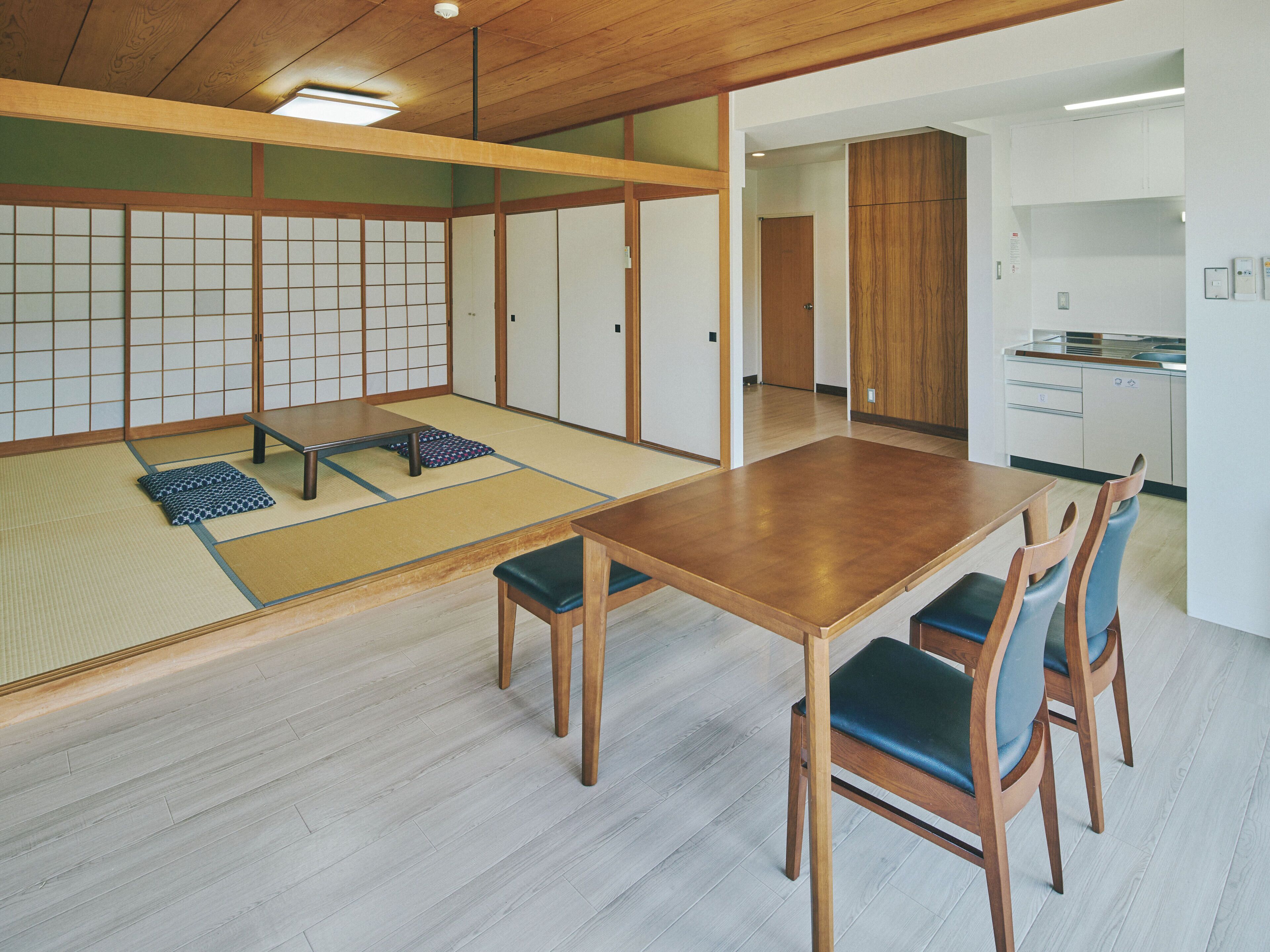 A traditional Japanese room with tatami flooring, a low wooden table, and a kitchen area visible through an open doorway.