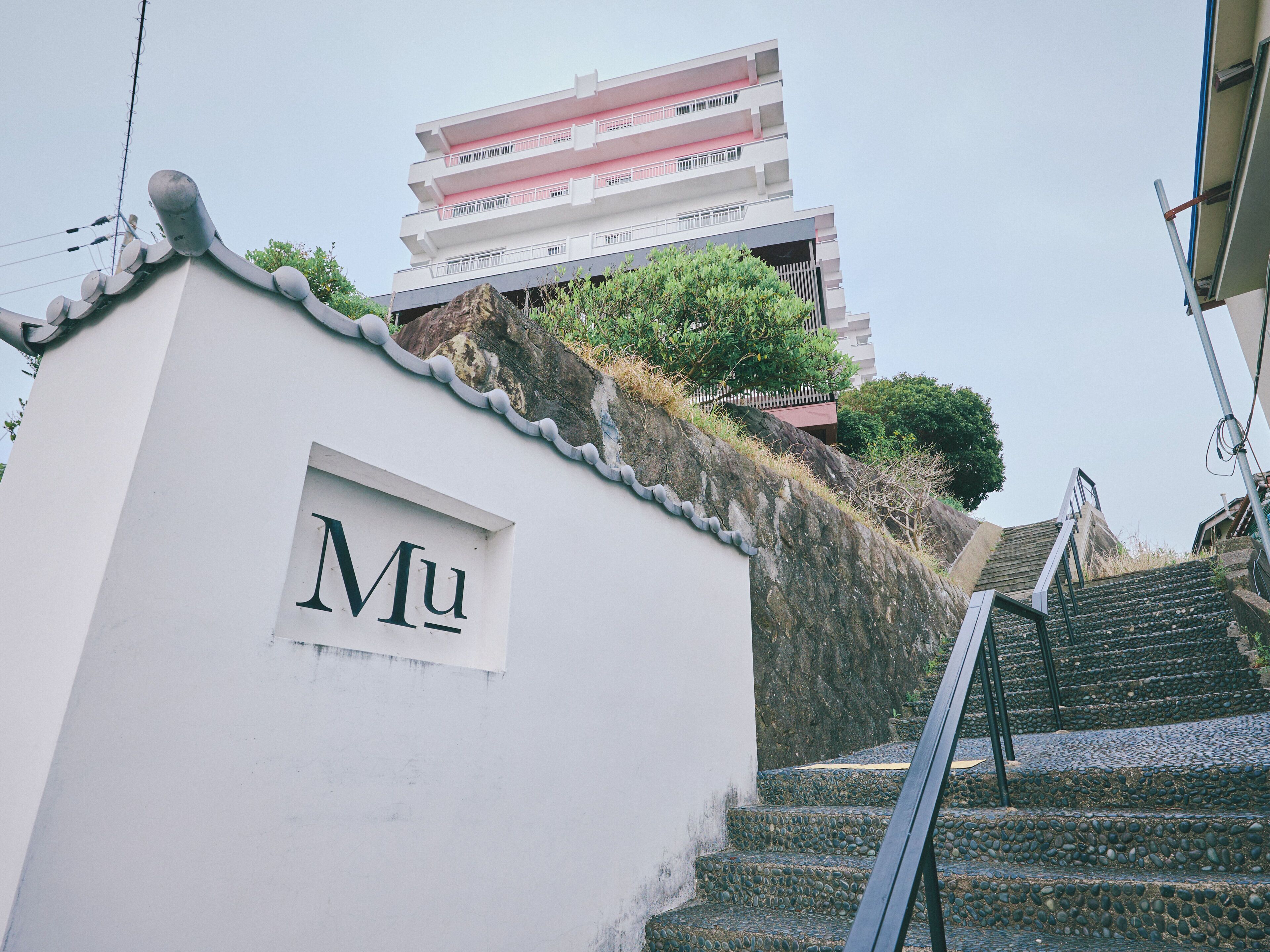 A white building with a sign reading 'Mu' and a staircase leading up to a multi-story building with balconies.