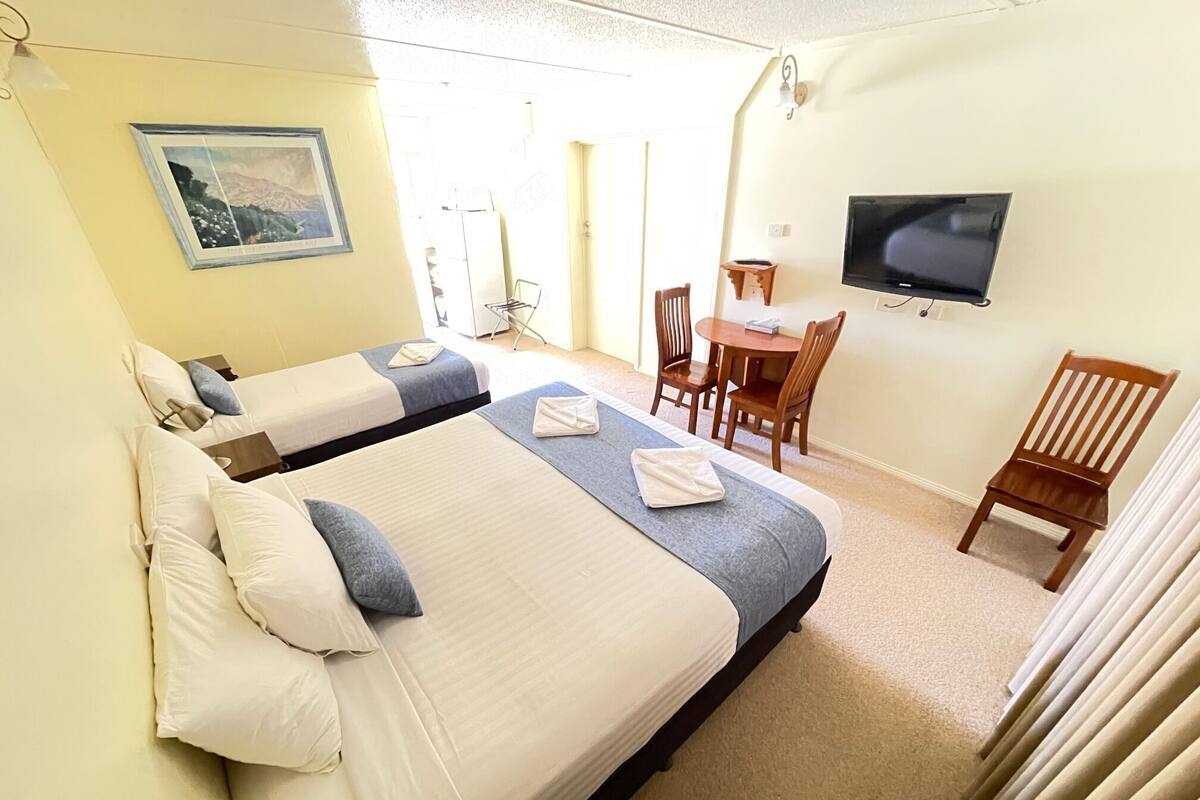 A hotel room with two beds, a dining table, chairs, a TV, and a painting on the wall.