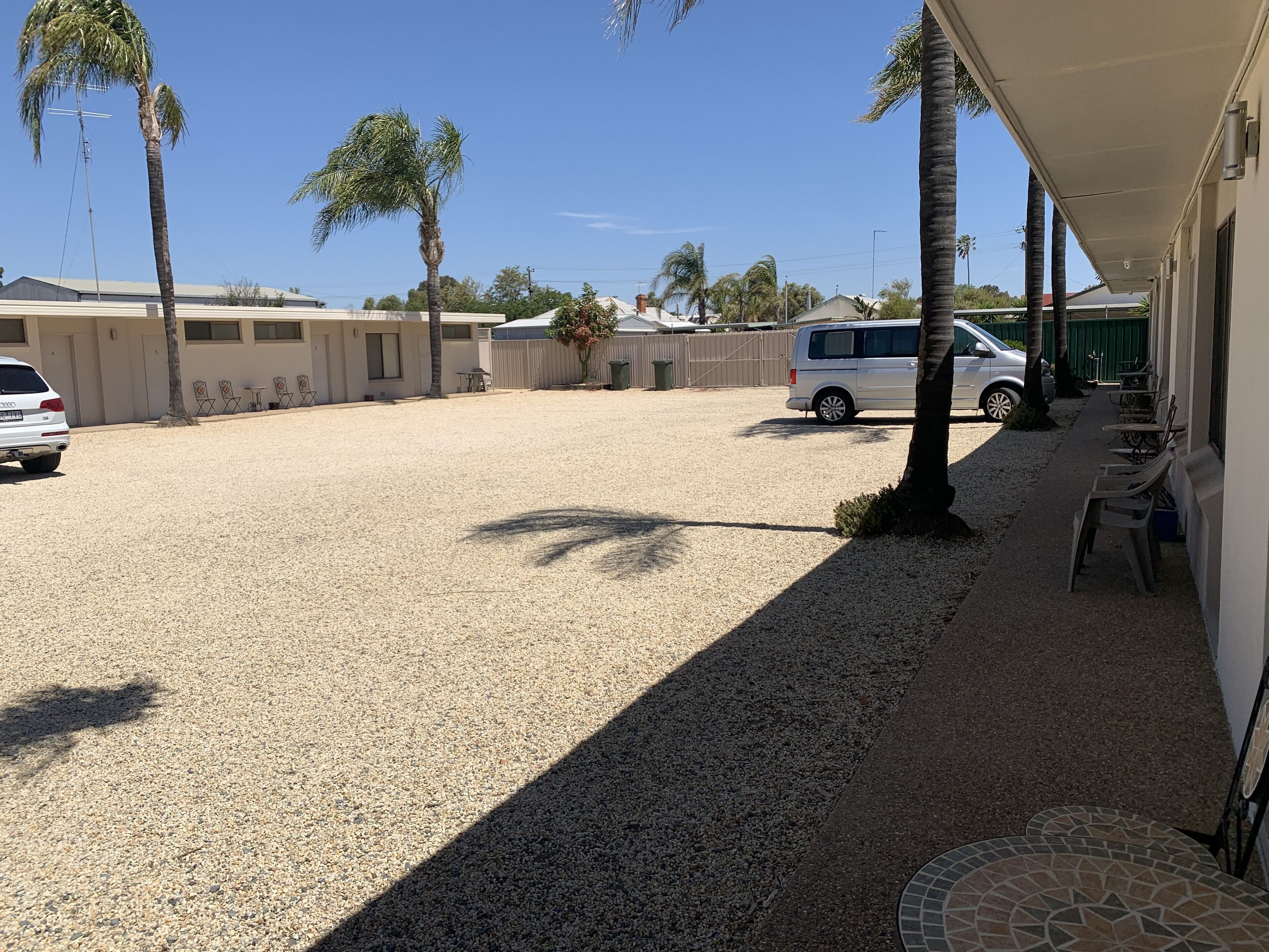 A covered outdoor area with a gravel ground, palm trees, a white van, and a building with a covered patio.