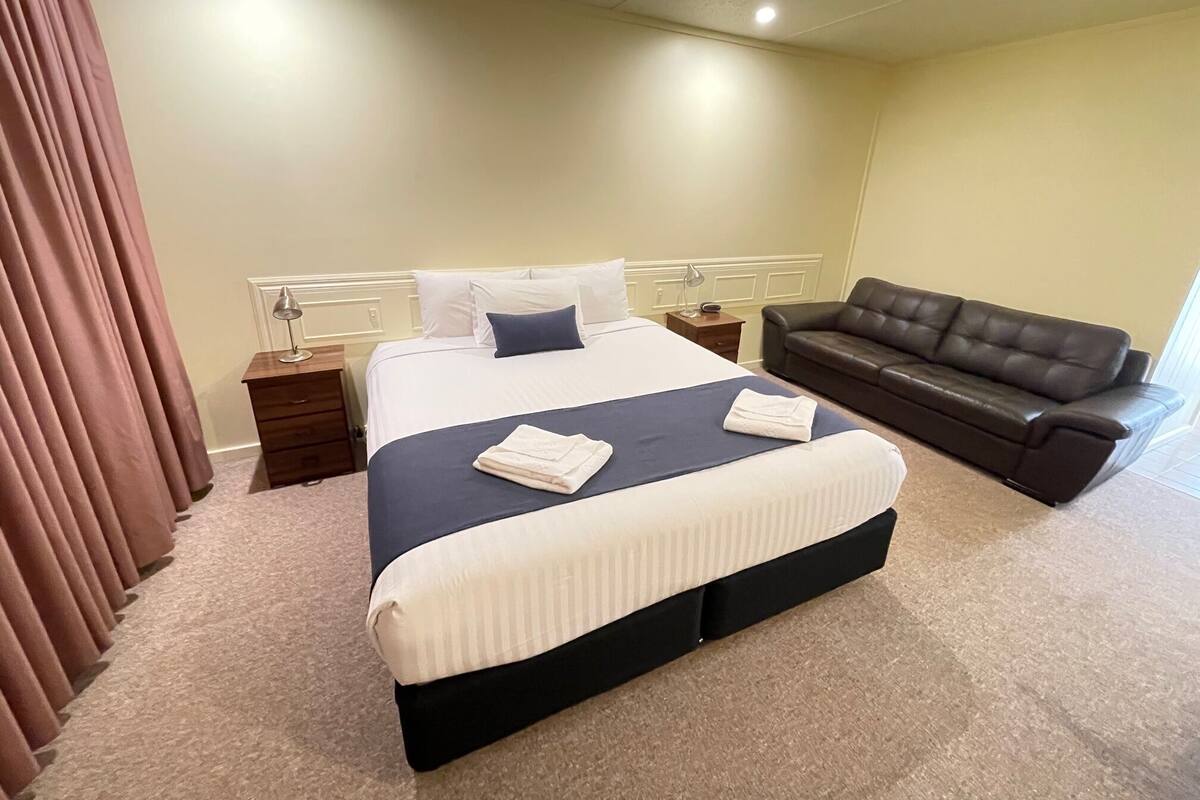 A hotel room with a bed, a nightstand, a lamp, a sofa, and a curtain.