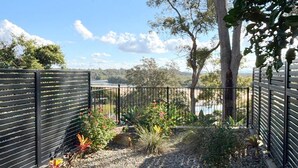 A garden with a metal fence, various plants, and a view of a body of water. - Nineteen On Gregory G02 - Slow M'Ocean - Linen Supplied (SOUTH WEST ROCKS)