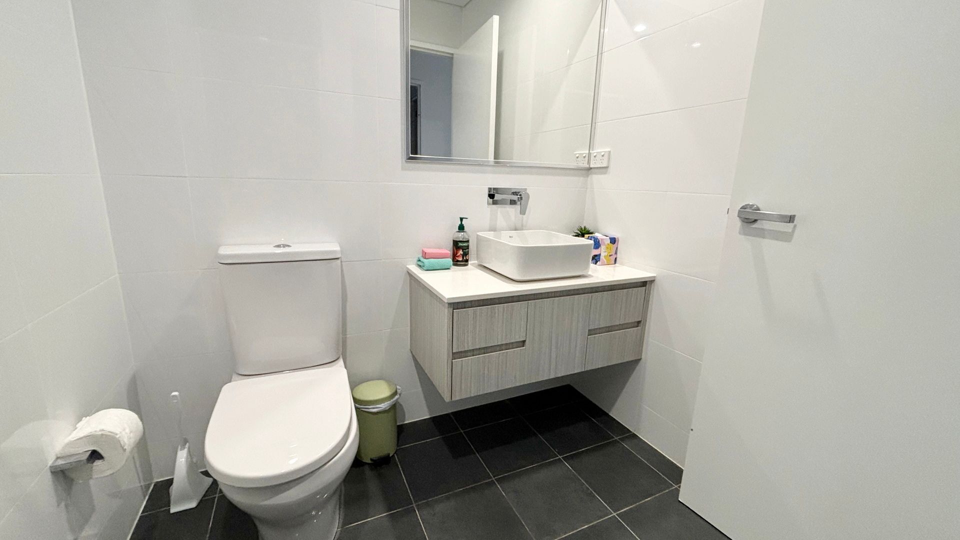 A bathroom with a toilet, a sink, a mirror, and a trash bin.