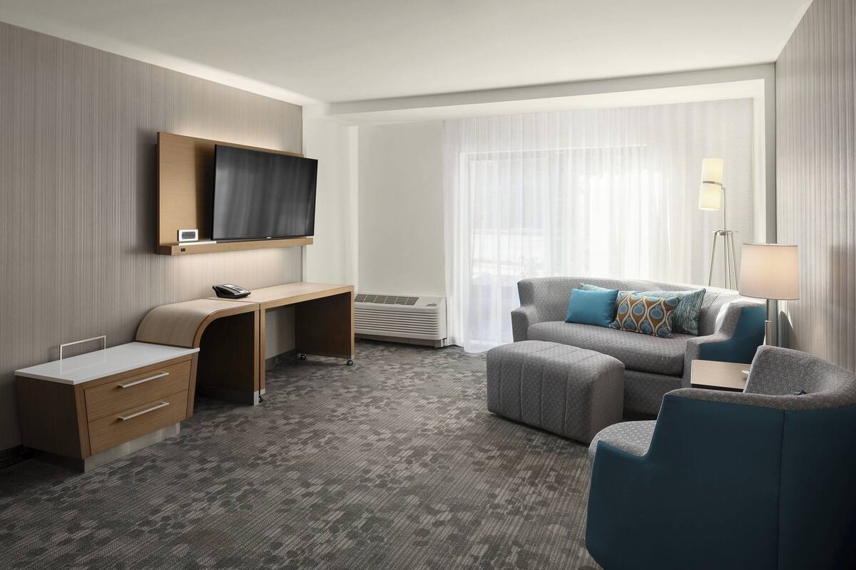 A hotel room with a flat-screen TV mounted on the wall, a desk with a computer, a sofa, and a chair.