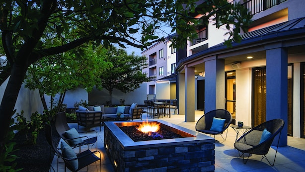 A modern outdoor patio with a stone fire pit, seating, and a view of a multi-story building.