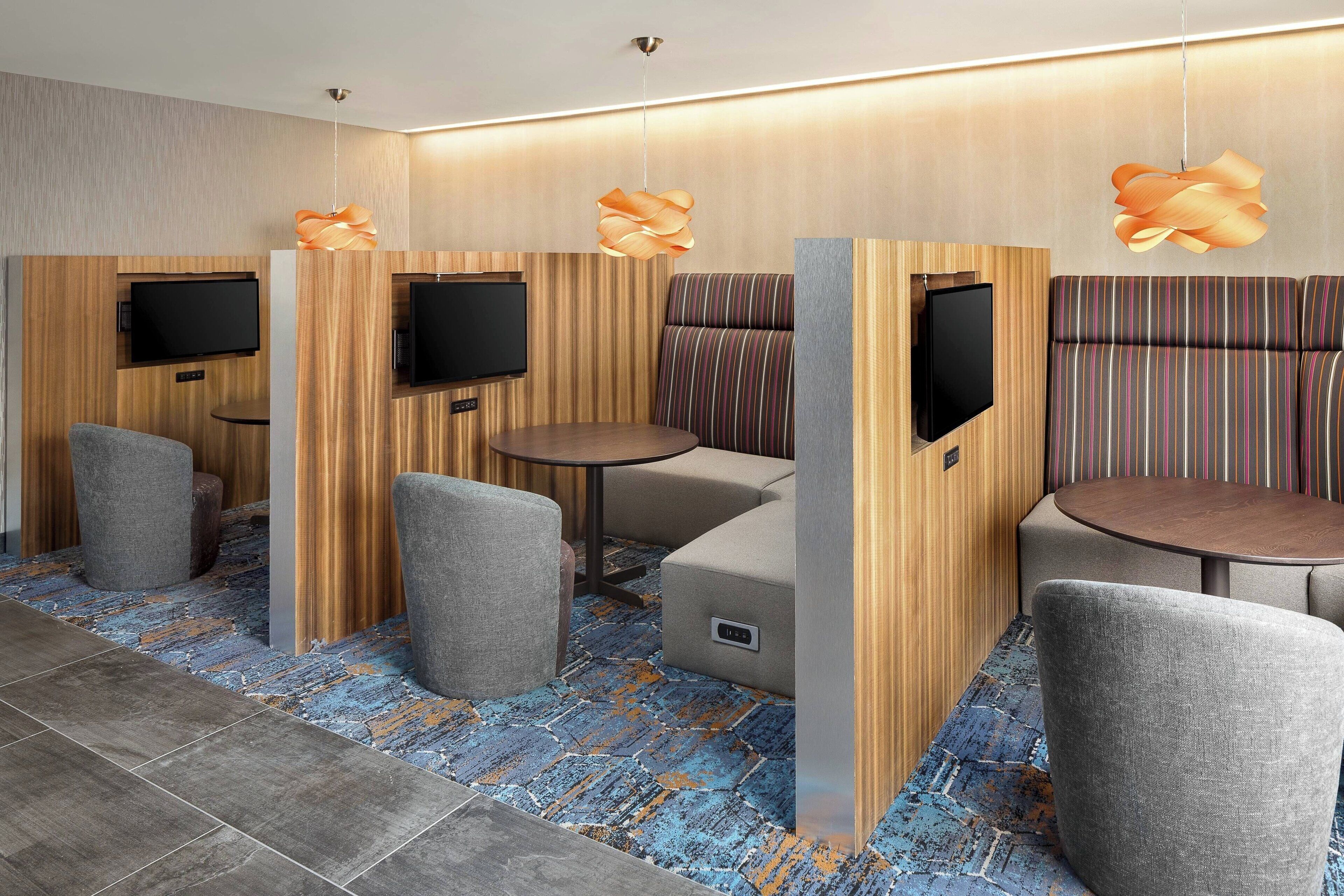 Quarto de hotel moderno com painel de parede em madeira, carpete com padrão azul e laranja, e um espaço de estar com uma mesa redonda e duas poltronas.